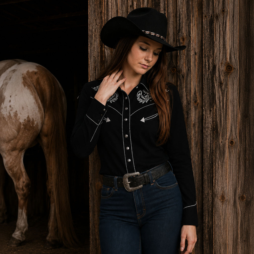 Woman wearing a black embroidered western cowgirl shirt with silver stallion detailing, dark cowboy hat, blue jeans, and a western belt beside a rustic barn and horse.