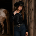 Woman wearing a black embroidered western cowgirl shirt with silver stallion detailing, dark cowboy hat, blue jeans, and a western belt beside a rustic barn and horse.
